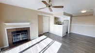 This single-floor space offers a clean, open-concept layout that seamlessly connects the living area to the kitchen. The living room is a welcoming, light-filled space, benefiting from natural sunlight that streams across the dark hardwood-style flooring. A ceiling fan with a classic brass fixture is centrally positioned overhead, providing both function and a traditional aesthetic touch. A tiled fireplace with a white mantel serves as a focal point along one wall, adding warmth and character to the room.

The kitchen is defined by crisp white cabinetry and a white peninsula that creates a natural divider between the cooking and living areas, also offering additional counter and seating space. The kitchen is equipped with a range and a full-size refrigerator, and features an overhead light fixture that keeps the space bright and functional.

The neutral warm-beige wall tones throughout complement the flooring and white trim, creating a cohesive and airy atmosphere. The space is finished with white baseboards and a clean white ceiling, contributing to its bright and well-maintained appearance. Overall, the layout is practical and inviting, with a straightforward flow that makes the space feel open and easy to move through.