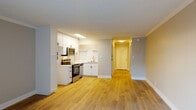 Located at 1738 Canyon Dr in Los Angeles, this single-floor unit offers a clean, modern aesthetic with thoughtful updates throughout. Light wood laminate flooring runs continuously across the entire space, creating a cohesive and inviting atmosphere. The open-concept living area is generously proportioned, featuring large sliding glass doors fitted with vertical blinds that invite abundant natural light and provide access to the outdoors. A wall-mounted air conditioning unit ensures year-round comfort. The kitchen is well-appointed with crisp white cabinetry extending to the ceiling for ample storage, stainless steel appliances including an electric range and built-in microwave, and a clean white countertop. A tall pantry cabinet adds additional storage. The hallway connecting the living areas features mirrored sliding closet doors on both sides, maximizing storage while enhancing the sense of space. A round overhead light fixture illuminates the corridor, which opens to additional rooms beyond. The bathroom is outfitted with a full-size soaking tub and shower combination in white, complemented by chrome fixtures and a matte black towel bar. Textured ceilings with crown molding details add a refined architectural touch throughout. The neutral gray and white color palette lends the space a fresh, contemporary feel throughout.