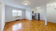 This single-floor space features a consistent design language anchored by warm parquet wood flooring laid in a classic basket-weave pattern throughout the living areas and bedrooms. The rooms are finished with crisp white walls, offering a bright, clean aesthetic that complements the natural warmth of the flooring.

The living area is generously proportioned, with a window providing natural light and views to the exterior. A baseboard heating unit and ceiling-mounted light fixture are present. The open sightline into the kitchen creates a connected feel between the two spaces.

The kitchen is equipped with warm-toned wooden cabinetry, a granite-look countertop with a stainless steel single-basin sink, a gas range with overhead ventilation, and a stainless steel refrigerator. Storage is provided by upper and lower cabinet configurations.

Additional rooms continue the parquet flooring theme and maintain the neutral white walls, with ceiling fixtures and doorway access to adjoining spaces. One room includes a closet, suitable for use as a bedroom.

The bathroom features classic black-and-white mosaic tile flooring, a white soaking tub with a shower rod and curtain track, a toilet, and a vanity with under-sink storage. A window above the tub allows for natural ventilation and light.