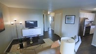 This single-floor space showcases a thoughtfully arranged living area that serves as the central hub of the unit. Dark hardwood flooring runs throughout, providing a rich contrast against the light, neutral-toned walls and white textured ceiling. The living room is furnished with a low-profile media console in crisp white, housing a flat-screen television, and is complemented by a rectangular coffee table atop a textured area rug, adding warmth and definition to the space. Framed artwork decorates the walls, contributing a personalized touch to the otherwise clean aesthetic. A floor lamp and table lamp offer layered lighting options throughout the room. A hallway visible from the living area leads to additional rooms, hinting at a well-connected floor plan. The corridor features a ceiling-mounted light fixture and access to what appears to be a closet and adjoining spaces beyond. Overall, the space presents a modern, minimalist design sensibility, with practical furnishings and a cohesive color palette that creates a comfortable and inviting atmosphere throughout.