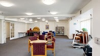 Located at 1745 E Fairway Dr in Orange, California, this single-story community facility offers a generous, well-organized layout across a substantial footprint. The space is designed to foster social interaction and comfortable daily living, with multiple distinct areas flowing together under a cohesive, light-filled interior.

The welcoming living room features plush blue sofas, warm wood accent tables, and large windows dressed with vertical blinds that allow ample natural light. A dedicated library and reading area showcases floor-to-ceiling bookshelves stocked with a wide collection, complemented by a round table and cushioned seating — ideal for quiet study or group discussion. An additional activity room features an upright piano, display shelving, and versatile seating arrangements suited for recreation or gatherings.

The open common area serves as a central hub, with multiple seating clusters and clear sightlines toward the entry, where a welcoming arrangement of chairs and a cheerful décor set a friendly tone. The functional kitchen is equipped with white cabinetry, a gas range, microwave, and full-size refrigerator, supporting meal preparation and community dining. A dedicated office rounds out the program, supporting administrative functions. Throughout, neutral carpet flooring, flush-mount ceiling lighting, and clean white walls create a calm, organized, and accessible environment.