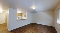 This single-floor space features a bright, open layout designed for functional living. The main living area is finished with rich, dark hardwood flooring that contrasts with the crisp white walls, creating a clean and inviting atmosphere. A flush-mount ceiling light provides overhead illumination, keeping the space feeling airy and well-lit.

A defining architectural feature is the pass-through window that connects the living area to the kitchen, promoting an open flow between the two spaces. The kitchen is equipped with light wood cabinetry, a microwave, a sink, and white appliances, all efficiently arranged within a compact footprint. The pass-through counter also serves as a convenient serving or casual dining surface.

The white walls throughout provide a neutral backdrop that enhances the sense of openness, while the white baseboards offer a tidy finishing detail. The overall design is straightforward and practical, with an emphasis on clean lines and efficient use of space. The layout is well-suited for those seeking a low-maintenance, move-in ready environment with a cohesive aesthetic throughout.