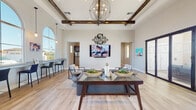 The Silvergate clubhouse in Brentwood, California is a thoughtfully designed single-story community space that blends rustic and contemporary elements to create a welcoming environment. The main gathering area immediately captures attention with its soaring ceilings adorned with rich, dark exposed wood beam detailing and a statement orb chandelier that anchors the room. Light wood-toned flooring flows throughout the entire space, complementing the crisp white walls and creating a bright, cohesive aesthetic.

Large arched windows along one wall flood the lounge area with abundant natural light, while floor-to-ceiling glass doors on the opposite side open directly to an outdoor patio and pool area, seamlessly connecting the interior to the exterior amenities. The lounge is furnished with comfortable seating arrangements centered around a glass-top coffee table, ideal for casual gatherings.

Adjacent to the main lounge, a fully equipped kitchen features dark espresso cabinetry, white subway tile backsplash, light quartz countertops, and stainless steel appliances. A built-in bar-height counter with seating provides a convenient casual dining or workspace area, illuminated by warm pendant lighting. The overall ambiance is modern, airy, and inviting, designed to serve as a functional and stylish hub for community residents.