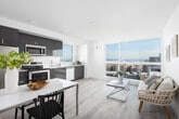 This contemporary single-floor residence showcases a clean, minimalist design defined by crisp white walls, light wood-style flooring, and an abundance of natural light throughout. The open-concept layout connects the kitchen, dining, and living areas into one cohesive space, making the interior feel airy and expansive.

The kitchen is a standout feature, outfitted with high-gloss gray cabinetry, stainless steel appliances including a refrigerator, range, microwave, and dishwasher, as well as a white countertop and a window that fills the space with natural light. A compact dining area sits adjacent, offering a seamless transition between cooking and entertaining.

The living area is bathed in light from large floor-to-ceiling windows that frame urban views of the surrounding cityscape, creating a bright and inviting atmosphere. The bedroom continues the light-filled aesthetic, highlighted by an oversized picture window that offers generous views of the exterior streetscape and sky.

A long, well-lit hallway connects the private areas of the residence, maintaining the home's clean sightlines and sense of openness. The bathroom features a modern vanity with a white countertop, a round mirror, gray cabinetry, and light wood-style flooring, rounding out the space with functional elegance. Throughout, the design prioritizes simplicity, light, and contemporary comfort.
