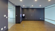 This contemporary single-floor space is defined by a clean, modern aesthetic that balances functionality with sleek design. The interior is characterized by deep navy-blue walls that create a bold, professional atmosphere throughout. Warm-toned hardwood flooring runs seamlessly across the space, providing an inviting contrast to the cool wall tones and light gray ceilings.

A standout architectural feature is the floor-to-ceiling glass wall system, which floods the space with abundant natural light and offers elevated panoramic views of the surrounding cityscape, hinting at a high-floor location within a larger building. Recessed ceiling lighting complements the natural light, ensuring the space is well-illuminated at all times.

The main area is equipped with a wall-mounted flat-screen television, making it well-suited for presentations, screenings, or collaborative gatherings. A wall-mounted digital control panel is also present, offering integrated building or room management capabilities. Multiple electrical outlets are distributed throughout the walls, supporting a variety of setups and configurations.

Glass partition walls further enhance the open, airy feel while allowing for visual connectivity. The overall ambiance is polished and versatile, lending itself to uses such as a meeting room, lounge, media room, or flexible event space.