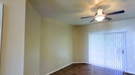 This single-story space presents a clean, functional layout highlighted by a combination of practical and comfortable design elements. The main living area features warm, light yellow-toned walls that create an inviting atmosphere, complemented by dark hardwood-style flooring that adds a grounding contrast to the room's soft color palette. White baseboards provide a crisp finish along the perimeter of the room.

A ceiling fan with an integrated light fixture is centrally positioned overhead, offering both illumination and air circulation throughout the space. The ceiling itself has a subtle textured finish, adding a modest layer of visual interest above.

Natural light is a notable feature of this room, introduced through a full-height sliding glass door fitted with vertical blinds. The door provides direct access to an outdoor area and, when the blinds are open, allows views of surrounding greenery, contributing to a bright and airy feel. The overall ambiance of the space is neutral and adaptable, making it well-suited for a variety of furnishing arrangements and interior styles.