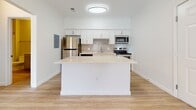 Located at 1100 S Missouri Ave in Clearwater, Florida, this single-floor unit offers a clean, modern design highlighted by light wood-style flooring and a bright, neutral color palette throughout. The open-concept layout centers around a well-appointed kitchen featuring white shaker cabinetry, a neutral tile backsplash, light quartz countertops, and a full suite of stainless steel appliances including a refrigerator, electric range, microwave, and dishwasher. A peninsula island provides additional prep space and naturally defines the transition into the open living area, which benefits from recessed and flush-mount lighting as well as crown molding details. The living space is generously proportioned and filled with natural light, with a tray ceiling adding architectural interest. Sliding glass doors lead to a private balcony overlooking neighboring rooftops and swaying palm trees, bringing a distinctly Florida ambiance to the space. A secondary bedroom features a large window with similar outdoor views. The bathroom is finished with a full soaking tub and shower combination set against white tile, complemented by a vanity with ample storage. Crown molding, consistent flooring, and a cohesive light color scheme unify the space, creating a calm and contemporary atmosphere throughout.