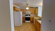 This single-floor space includes a well-equipped kitchen designed for everyday functionality. The kitchen features warm-toned, light wood cabinetry that extends both above and below the counter, providing generous storage throughout. Upper cabinets line the walls, maximizing vertical space, while lower cabinets and drawers offer additional organization options beneath the countertops.

The kitchen is outfitted with a full suite of white appliances, including a refrigerator, an electric range with oven, an over-the-range microwave, and a double-basin stainless steel sink positioned along the side wall. The layout follows a U-shaped configuration, creating an efficient workflow between the primary cooking and prep areas.

A ceiling fan with an integrated light fixture is centrally mounted, ensuring comfortable air circulation and supplemental lighting throughout the space. The floor features durable tile in warm earth tones, complementing the cabinetry and contributing to the kitchen's cohesive, neutral palette. The textured ceiling adds a subtle visual element overhead. Overall, the kitchen presents a practical and tidy environment well-suited for daily use.