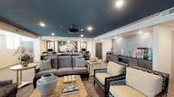 This single-floor amenity space is designed around a cinematic theme, combining screening, lounge, and business functions in one cohesive environment. A bold teal-painted ceiling anchors the room's design, complemented by recessed lighting and a ceiling-mounted projector that serves a large built-in projection screen framed by ambient LED lighting. Classic movie posters and film reel artwork decorate the walls, reinforcing the theatrical atmosphere throughout.

The main lounge area features a generously sized gray sectional sofa and armchairs upholstered in teal and navy chevron and geometric patterns, arranged around a rustic wood coffee table. A pair of deep wingback chairs provides additional seating near the screening wall, creating an intimate viewing setup. Decorative accessories, including ceramic vases and floral arrangements, add refined touches throughout.

Along one wall, a wet bar station with dark cabinetry and a glittering mosaic tile backsplash is equipped for entertaining, with a popcorn machine and glassware on display. Adjacent to this, a casual dining area offers round tables with patterned upholstered chairs. A dedicated business corner includes computer workstations and a desk area. Patterned carpeting, neutral walls, and small windows completing the well-appointed, multifunctional space.
