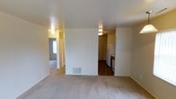 This single-floor residential unit offers a straightforward and functional layout designed for comfortable everyday living. The main living area is finished with light-toned carpet, providing a warm and inviting atmosphere. A floor-mounted HVAC vent is integrated into the baseboard, ensuring year-round climate comfort throughout the space.

The living area opens directly toward the kitchen, which is distinguished by dark hardwood-style flooring that creates a clear visual separation between the two spaces. The kitchen is equipped with a white refrigerator and a stove, and features dark cabinetry that adds contrast to the light-colored walls. Flush-mounted ceiling light fixtures provide practical illumination throughout both areas.

A hallway visible from the living area leads to additional rooms, suggesting a multi-room configuration that includes further private spaces. The walls throughout are painted in a clean, neutral white, contributing to a bright and airy feel. The open sightlines between the living area, kitchen, and hallway enhance the sense of connectivity and flow within the unit. Overall, the space presents a clean, move-in-ready environment with a simple, utilitarian design aesthetic suited for a variety of living arrangements.