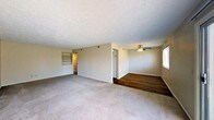 This single-floor residence offers a straightforward and functional layout designed for comfortable everyday living. The main living area is generously proportioned, featuring neutral-toned wall-to-wall carpeting that creates a warm and inviting atmosphere. A textured popcorn ceiling adds a classic finish throughout the space, while a ceiling fan in the adjoining area provides both airflow and ambient lighting.

The open layout connects the main living zone to a secondary area distinguished by rich, dark hardwood flooring, offering a natural contrast to the carpeted spaces. Built-in shelving along one wall provides convenient storage and display options, adding a practical architectural detail to the living area.

Natural light filters in through a window along the side wall, brightening the interior and enhancing the sense of openness. Neutral walls throughout keep the space versatile and move-in ready. A glimpse into the adjacent areas reveals a refrigerator, suggesting a kitchen nearby, as well as a hallway leading further into the unit. The overall ambiance is clean, understated, and functional, making it a practical canvas ready to be personalized to suit a variety of lifestyles.