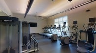 This contemporary fitness center is thoughtfully designed to provide a comprehensive workout environment on a single level. The space features a sleek, modern aesthetic with a neutral color palette, dark rubberized flooring, and a coffered ceiling with warm recessed lighting accented by exposed industrial-style beams and ceiling fans for optimal air circulation.

The cardio area is well-stocked with a row of treadmills aligned along the wall, complemented by elliptical trainers and additional cardio equipment, all from Precor — a leading commercial fitness brand. A wall-mounted flat-screen television is positioned for convenient viewing during workouts. Strength and resistance training equipment is available on the opposite side of the floor, rounding out the exercise offerings.

Natural light flows into the space through a large window fitted with roller shades, offering both brightness and the option for glare control. The open layout ensures ample room to move between stations comfortably, creating an uncluttered and functional training environment. The overall ambiance is clean, professional, and motivating — well-suited for daily fitness routines.