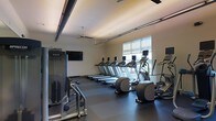 This contemporary fitness center is thoughtfully designed to provide a comprehensive workout environment on a single level. The space features a sleek, modern aesthetic with a neutral color palette, dark rubberized flooring, and a coffered ceiling with warm recessed lighting accented by exposed industrial-style beams and ceiling fans for optimal air circulation.

The cardio area is well-stocked with a row of treadmills aligned along the wall, complemented by elliptical trainers and additional cardio equipment, all from Precor — a leading commercial fitness brand. A wall-mounted flat-screen television is positioned for convenient viewing during workouts. Strength and resistance training equipment is available on the opposite side of the floor, rounding out the exercise offerings.

Natural light flows into the space through a large window fitted with roller shades, offering both brightness and the option for glare control. The open layout ensures ample room to move between stations comfortably, creating an uncluttered and functional training environment. The overall ambiance is clean, professional, and motivating — well-suited for daily fitness routines.