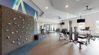 This fitness center in West Melbourne, Florida, offers a comprehensive workout environment designed to accommodate a wide range of exercise needs. The single-floor facility features a modern, open layout with recessed lighting, tray ceilings, and ceiling fans that contribute to a clean, airy atmosphere throughout.

A standout architectural element is the bold "Freedom Climber" rock climbing wall, set against a vibrant graphic mural with geometric mountain-inspired designs in blue, teal, and yellow. This feature anchors one end of the space and adds a distinctive visual focal point. Rubber flooring in complementary tones covers the workout areas, providing both safety and durability.

The strength training zone includes a power rack with free weights, a multi-station cable machine, and a full dumbbell rack. A large floor-to-ceiling mirror expands the visual space and supports proper form during workouts. The cardio section is lined with treadmills, rowing machines, and specialty bikes, accompanied by ceiling-mounted televisions for entertainment.

A glass panel garage door opens to connect an interior workout bay to the outdoors, flooding the space with natural light and offering views of a lush green outdoor area with palm trees. Additional windows throughout provide further natural light and ventilation, enhancing the overall workout environment.