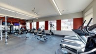This fitness center offers a comprehensive workout environment designed to accommodate a wide range of exercise routines. The space is finished with durable dark rubber flooring, providing both safety and functionality for various training activities. Large horizontal windows line one wall, framed by bold red accent panels that add a striking visual contrast to the otherwise neutral white and gray palette. These windows allow ample natural light to fill the room while offering views of the surrounding outdoor environment.

Overhead, linear LED lighting runs along a tiered ceiling, supplemented by a ceiling fan that aids in air circulation throughout the space. The layout is thoughtfully organized, with cardio equipment — including multiple treadmills — positioned along one side, and a dedicated strength training area on the other. The strength zone includes a cable machine, weight bench, and a power rack with free weights and medicine balls, making it suitable for both beginner and advanced training. A mirrored wall near the free weights section enhances spatial awareness during workouts. The overall design is clean, modern, and functional.