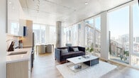 This contemporary urban residence showcases an industrial-modern aesthetic defined by exposed concrete ceilings, light hardwood flooring, and an abundance of natural light. The open-concept living and dining area is anchored by dramatic floor-to-ceiling windows on multiple walls, framing expansive city skyline views and flooding the space with daylight. Structural concrete columns add architectural character to the living area, which comfortably accommodates both a lounge and dining setup.

The kitchen is thoughtfully designed with high-gloss white upper cabinetry, light wood lower cabinets, quartz countertops, a white subway tile backsplash, and a full suite of stainless steel appliances — combining sleek style with practical functionality.

A clean, light-filled hallway with an exposed concrete ceiling connects the living spaces to the private quarters. The bedrooms feature the same warm hardwood floors, soaring ceilings, and access to natural light, with one bedroom benefiting from a large window with city views. Closet space is integrated into the bedroom layout for added convenience.

The bathroom is finished with large-format linear tile in the walk-in glass shower, mosaic tile flooring, and polished chrome fixtures, conveying a spa-like refinement. Throughout the residence, the interplay of raw concrete, clean white walls, and warm wood tones creates a sophisticated, urban atmosphere.