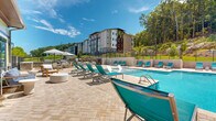 The pool area at Canopy Apartment Villas in Pelham, Alabama offers a well-appointed outdoor amenity space designed for relaxation and recreation. The centerpiece is a generously sized swimming pool with crystal-clear blue water, complemented by a separate spa or wading feature along its edge. In-pool lounge chairs allow residents to relax directly in the water, while a broad paver deck surrounds the pool on all sides, providing ample space for sunbathing.

Teal-colored chaise lounges are arranged throughout the deck, along with shaded seating areas featuring market umbrellas and casual furniture groupings. The deck's herringbone-patterned paver design adds a refined, resort-like aesthetic to the space.

Adjacent to the pool is a striking covered pavilion structure, distinguished by exposed heavy timber framing, steeply pitched rooflines, and stacked stone columns with flat-top caps. Large ceiling fans keep the covered area comfortable, and the open-air design allows for natural ventilation. The pavilion connects to an interior amenity building with large glass doors.

The pool area is enclosed by a black iron fence, with a natural wooded hillside providing a lush, private backdrop. The multi-story residential buildings of Canopy Apartment Villas are visible nearby, framing the outdoor space within the broader community setting.