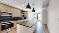 Located at 1300 40th St in Denver, CO, this modern one-bedroom, one-bathroom unit offers a thoughtfully designed open-concept layout with contemporary finishes throughout. Light wood-tone flooring flows seamlessly through the main living areas, complementing the clean, neutral palette of the space.

The kitchen serves as the heart of the unit, featuring flat-front light wood cabinetry, dark subway tile backsplash, a dark quartz countertop island with pendant lighting overhead, and a full suite of stainless steel appliances including a gas range, refrigerator, microwave, and dishwasher. Open shelving adds a modern, functional touch.

The kitchen opens directly into the living area, which is bright and airy thanks to large windows and a glass door leading to an outdoor balcony with views of the Denver skyline. The bedroom offers plush carpeting, a large window for natural light, and direct access to the balcony as well.

The bathroom is well-appointed with light wood cabinetry, a dark quartz vanity countertop, black hardware accents, and wood-look flooring that ties the space together. A dedicated closet is accessible from the bathroom, providing convenient storage. The overall design aesthetic is clean, contemporary, and well-executed.