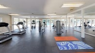 This well-equipped fitness center at 1401 E Millbrook Rd in Raleigh, NC offers a comprehensive workout environment designed to support a wide range of exercise routines. The single-floor facility is organized into distinct zones, providing ample space for both cardio training and strength work without feeling crowded.

The cardio section features multiple treadmills, elliptical trainers, and a rowing machine, giving users a variety of aerobic options. Alongside these, a selection of strength and resistance machines rounds out the equipment offerings for a full-body workout experience.

A dedicated stretching and functional training area is clearly defined by colorful exercise mats laid across the durable rubberized flooring, which spans the entire facility. Large wall mirrors in this zone enhance spatial awareness during functional movements and accessory exercises, while medicine balls and other small training tools are conveniently available nearby.

Natural light fills the space through an extensive bank of windows and glass doors that line one wall, offering pleasant views of an outdoor pool area. White structural columns punctuate the open floor plan, and ceiling fans help maintain a comfortable environment throughout. Overhead fluorescent lighting ensures the space remains bright at all hours. The overall ambiance is clean, functional, and inviting — well-suited for daily fitness use.