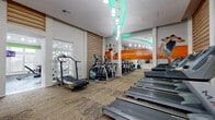 This modern fitness center is thoughtfully designed across a single floor, offering a comprehensive workout environment divided into distinct functional zones. The cardio area features a row of treadmills, elliptical machines, and stationary bikes — including upright and recumbent models — set atop patterned carpet tiles. An eye-catching teal LED curved accent on the ceiling, paired with elegant pendant lighting, gives this zone a dynamic, energizing ambiance. Full-length mirrors line the walls and are complemented by bold orange motivational wall graphics, amplifying the sense of space and inspiring movement. Adjacent to the cardio zone, a dedicated strength training area offers rubber flooring, weight benches, cable machines, selectorized strength equipment, and a dumbbell rack. This section benefits from soaring ceilings, recessed lighting, and a bright color-blocked green accent wall, with large windows fitted with plantation shutters allowing natural light to fill the room. A separate open-plan area with hardwood flooring provides space for stretching, group cycling, or functional training. Decorative perforated white panels and warm wood slatted wall accents add architectural interest throughout, creating a polished, boutique-gym aesthetic that is both functional and visually distinctive.