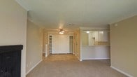 This single-story space offers an open and functional layout that connects the main living area to the kitchen. The living room is finished with soft, neutral-toned carpet that contributes to a warm and comfortable atmosphere. Crown molding runs along the perimeter of the ceiling, adding a classic architectural detail, while a ceiling fan with a light fixture serves as the central overhead feature.

A built-in shelving unit is positioned along one wall, providing convenient display and storage space, while a set of sliding closet doors nearby offers additional storage. A decorative column subtly defines the transition between areas, lending a sense of structure to the open floor plan.

The kitchen is accessible via a pass-through counter, which opens the space visually and allows for easy interaction between the kitchen and living areas. White cabinetry lines the kitchen walls, providing ample storage, and the countertop extends to form a practical breakfast bar. Tile flooring in the kitchen contrasts neatly with the carpeted living space. The textured ceiling and consistent neutral palette tie all areas of the space together cohesively.
