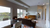 This single-floor space at Bishops offers a well-appointed living area characterized by a neutral color palette with warm taupe and gray tones on the walls, creating a calm and inviting atmosphere. The living room is generously sized, anchored by a large, plush brown sofa and complemented by a bold floral area rug in blue, orange, and white that adds a striking visual focal point to the space. An orange accent chair introduces a pop of color, while a carved wooden sideboard adds an element of artisanal craftsmanship to the room. Natural light filters through large double-hung windows, offering views of the surrounding outdoor landscape and contributing to an airy, open feel. Recessed ceiling details add architectural interest overhead, while contemporary floor lamps provide warm supplemental lighting. A glass-top dining or accent table is visible in the foreground, reflecting light and adding a modern touch. An adjacent room is partially visible, hinting at additional living space beyond. Overall, the space balances contemporary design with eclectic furnishing choices for a comfortable, stylish environment.