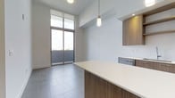 This single-floor space showcases a clean, contemporary design with an open-plan layout connecting the kitchen and living areas. The kitchen features a generous island with a crisp white countertop and warm wood-toned cabinetry that extends to the upper cabinets, providing ample storage. A built-in stainless steel appliance is seamlessly integrated beneath the counter, contributing to the sleek aesthetic. A stylish cylindrical pendant light hangs above the island, adding a refined touch to the space.

The living area benefits from impressively tall ceilings that create a sense of openness and volume. Large floor-to-ceiling sliding glass doors flood the interior with abundant natural light and offer direct access to an outdoor balcony, framing views of the surrounding environment. The large-format grey tile flooring runs consistently throughout, unifying the space and complementing the neutral white walls. Roller shades on the windows allow for adjustable light control. Overall, the space presents a bright, airy, and well-appointed environment with a modern minimalist character.