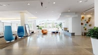 This single-floor commercial space presents a striking first impression through its expansive lobby, defined by large-format polished tile flooring that reflects an abundance of natural light streaming in through floor-to-ceiling glass windows and sliding doors. The open-plan lobby features a clean, contemporary design with crisp white walls and recessed ceiling lighting. A built-in fireplace anchors one side of the room, while a curated seating arrangement — including a vibrant orange lounge chair, a circular area rug, and a navy ottoman — creates an inviting focal point at the center of the space. Lush potted plants are positioned throughout, adding a natural, refreshing contrast to the minimalist aesthetic. Beyond the lobby, the space transitions into a more intimate reception area featuring rich dark wood cabinetry, patterned upholstered guest chairs, and dark-toned carpeting that lends warmth and a sense of professionalism. A dedicated reception desk serves as the functional hub of this area, flanked by decorative greenery including tall dracaena and snake plants. The overall ambiance throughout the space balances modern sophistication with approachable comfort, making it well-suited for a professional office or service-oriented environment.