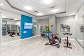 This contemporary fitness center is thoughtfully designed to support a full range of workout routines across several distinct zones. The main cardio area features a generous selection of treadmills, ellipticals, and stair climbers, each equipped with individual display screens. The space is anchored by a striking open-grid ceiling with decorative pendant lighting, and the richly patterned carpet in deep brown and teal tones adds a polished, upscale feel throughout.

A dedicated cycling and group fitness studio is separated from the main floor by glass partition walls, offering a bright and open atmosphere with light hardwood-style flooring. This area is equipped with stationary spin bikes and an array of colorful medicine balls and resistance tools neatly organized on wall-mounted racks. A large flat-screen display and floor-to-ceiling mirrors enhance the studio experience.

The functional training area includes a multi-cable rig, rowing machine, plyo box, and additional strength machines, all set against large windows that invite natural light. Mirrored walls throughout the facility create a sense of expansive space. A glass-enclosed changing or restroom area with tile flooring provides convenient amenities. The overall design blends modern aesthetics with practical functionality, creating a professional and motivating environment for all fitness levels.