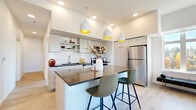 Located at 10306 SE Main St in Milwaukie, Oregon, this well-designed two-bedroom, two-bathroom residence offers a thoughtful single-level layout with clean, contemporary finishes throughout. The open-concept kitchen and living area serves as the heart of the home, where a striking island with a dark countertop and seating for two anchors the space. Pendant lights with warm gold interiors hang above, complementing white cabinetry, a subway tile backsplash, and stainless steel appliances. The adjacent living room benefits from a large picture window that invites generous natural light and views of the surrounding neighborhood. Light wood-tone flooring flows consistently throughout, creating a cohesive and airy aesthetic. The primary bedroom is a comfortable retreat, featuring ample natural light from wide windows and durable wood-look flooring. The en-suite bathroom offers a dark-countered vanity, white cabinetry, and a glass-enclosed walk-in shower with subway tile surround. The second bedroom includes a sliding-door closet, while the second bathroom mirrors the same refined, minimalist style. A dedicated laundry room with stacked washer and dryer adds everyday convenience. Hallways are well-proportioned and decorated tastefully, completing this polished, move-in-ready home.