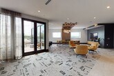 This contemporary amenity lounge is thoughtfully designed to serve as a versatile gathering and relaxation space. The open floor plan flows seamlessly across a bold geometric-patterned carpet that anchors the entire area, creating a cohesive and visually dynamic foundation throughout.

A striking architectural detail draws the eye immediately: a sculptural arrangement of angled wood slats descends from the ceiling, complemented by a cascading cluster of globe pendant lights that cast a warm, ambient glow over the central dining table and upholstered seating. The dining zone features a wood-toned rectangular table surrounded by mustard-hued chairs, ideal for collaborative gatherings or casual meals.

The lounge area is furnished with plush golden leather armchairs and a comfortable grey sectional sofa accented with decorative pillows in emerald green — a rich contrast against the muted grey accent walls. A wall-mounted flat-screen television and a low wood media console anchor one end of the room, creating an inviting media and relaxation corner.

A built-in kitchenette with warm wood cabinetry, a beverage station, and under-counter refrigeration provides convenient amenities for residents or guests. Natural light fills the space through multiple horizontal windows, while oversized black-framed French doors open directly onto a furnished outdoor terrace, offering treetop and skyline views. Colorful abstract artwork adds curated artistic character to the space.