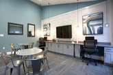 Located at 550 St. Michaels Way in Newport News, Virginia, this single-floor community clubroom offers a well-appointed and versatile gathering space designed for both relaxation and productivity. The room features soaring vaulted ceilings with a ceiling fan and recessed lighting, creating an open and airy atmosphere throughout. Walls are finished in a calming slate blue, complemented by crisp white millwork, textured wave-panel wall accents, and patterned carpet tile flooring that adds visual interest underfoot. Multiple round café-style tables with metal chairs are arranged throughout, providing flexible seating for socializing or casual meetings. A built-in workstation along one wall includes ample cabinetry, a mounted flat-screen television, computer monitors, and decorative pendant lighting, making it suitable for remote work or business use. Contemporary artwork adorns the walls, contributing to the polished aesthetic. Large black-framed windows and a glass sliding door flood the space with natural light while offering direct views of the outdoor pool area and surrounding community grounds. A framed wall mirror and a glass-panel interior door add additional character. The space is accessible via multiple entry points, including an exterior-facing door, making it a convenient and inviting hub for community residents.