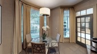 This refined single-floor space exudes understated elegance through a carefully curated palette of warm neutrals and sophisticated design details. The dining area serves as the heart of the room, centered around a circular glass-top table surrounded by woven rattan-style chairs, one of which features a richly patterned upholstered seat. A modern drum pendant light hangs above, casting a soft glow over the space. The walls are finished in a textured grasscloth-style wallcovering that adds depth and warmth to the overall atmosphere. Multiple large windows, dressed with floor-to-ceiling champagne-toned drapes and wood-slat blinds, flood the interior with natural light and offer views of lush greenery outside. A dark-framed glass-paned door with transom windows provides a striking architectural accent while connecting the space to an adjacent area. Crown molding runs along the ceiling, adding a classical finishing touch. A dark espresso-toned sideboard cabinet along one wall houses a beverage station and is topped with a serene abstract artwork illuminated by a picture light, contributing to the room's curated, gallery-like quality. The seamless blend of natural textures, refined furnishings, and ample natural light creates a warm yet polished ambiance ideal for intimate dining.