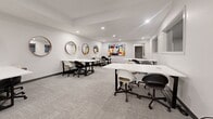Located at 125 Broad St in Weymouth, Massachusetts, this modern single-floor workspace offers a clean, contemporary environment designed to support a variety of professional needs. The interior is characterized by crisp white walls, recessed lighting, and neutral-toned carpet tiles that create a bright, focused atmosphere throughout.

The main open area features multiple white rectangular tables paired with a mix of ergonomic black and white rolling chairs, providing flexible seating arrangements suited for collaborative work, training sessions, or individual focus time. A series of round wood-framed mirrors lines the walls, adding a stylish design element and enhancing the sense of space. Bold, colorful artwork punctuates the white walls, contributing energy and creativity to the environment.

Adjacent to the main workspace, a smaller private meeting room offers a more intimate setting with its own table and seating, ideal for confidential discussions or small team gatherings. Interior windows between areas allow for visual connectivity while maintaining separation. A secondary meeting or breakout room near the entrance provides additional flexibility for private use.

The layout is thoughtfully organized, maximizing usable floor space while maintaining clear circulation paths. The space is well-suited for coworking, professional training, or small business operations, offering a polished and functional setting in the heart of Weymouth.