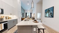Located at 505 West Eighth Street in Dallas, TX, this thoughtfully designed one-bedroom, one-bathroom residence offers an open-concept layout that seamlessly integrates the living, dining, and kitchen areas into one cohesive, airy space. Soaring ceilings amplify the sense of openness throughout, while light wood-tone flooring adds warmth and continuity underfoot.

The kitchen is a standout feature, showcasing two-tone cabinetry with soft gray lower cabinets and crisp white uppers, all accented with brushed gold hardware. A herringbone tile backsplash, quartz countertops, stainless steel appliances, and a large center island with seating for multiple guests make this kitchen as functional as it is elegant. Smoked glass pendant lights hang above the island, adding a refined touch. A dedicated pantry and in-unit laundry provide added convenience.

The living area is bathed in natural light from large windows overlooking the surroundings, creating a bright and welcoming atmosphere. The spacious primary bedroom continues the natural light theme with generous windows, soft carpet underfoot, and a teal accent wall that adds depth and character. A dedicated closet connects directly to the ensuite bathroom, which features white cabinetry with gold hardware, a glass-enclosed shower, and clean, minimal finishes throughout.