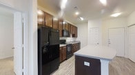This single-floor residence is designed with an open-concept layout that connects the kitchen, dining, and living areas into one cohesive and functional space. The kitchen is a standout feature, showcasing rich dark wood cabinetry that extends to the upper walls, providing generous storage throughout. A central island with a light granite-style countertop doubles as a breakfast bar, offering casual seating for everyday dining. Black appliances, including a range, microwave, and refrigerator, add a sleek, uniform look to the kitchen. The dining area flows naturally from the kitchen, making it ideal for entertaining. Recessed lighting and ceiling fans are installed throughout, ensuring both practical illumination and comfortable airflow. The living area, visible from the kitchen, features light-toned walls accented with coastal-inspired décor, creating a relaxed and welcoming ambiance. Light hardwood-style flooring runs throughout the main living spaces, contributing to a bright and airy atmosphere. Windows allow natural light to filter in, enhancing the open feel of the interior. Overall, this residence combines functional design with warm, modern finishes to create a comfortable and inviting living environment.