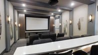This dedicated screening room delivers a true cinematic experience through thoughtful design and high-end finishes. At the focal point of the space sits a large, professionally framed projection screen, flanked by vertical wood slat panels that add warmth and acoustic depth to the otherwise contemporary aesthetic. Rows of plush, dark leather recliner-style seats are arranged in a tiered layout, ensuring unobstructed sightlines from every position in the room. Recessed ceiling lights and elegant wall sconces provide warm, ambient illumination that complements the theater's moody atmosphere without producing unwanted glare on the screen. Overhead, a ceiling-mounted projector anchors the audiovisual setup, while structured ceiling beams add an architectural dimension to the space. Carpeted flooring throughout aids in sound absorption, contributing to the room's acoustically considered environment. A sleek countertop surface near the rear of the room serves as a practical ledge, ideal for refreshments or equipment. The overall design balances functionality with sophistication, making this space equally suited for private movie screenings, presentations, or entertainment events.