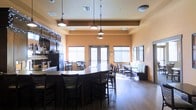 This single-floor space is designed to accommodate a variety of dining and social experiences. At its center sits a long, dark-finished bar counter surrounded by high-backed bar stools, creating a natural focal point for the room. Pendant lights hang at varying heights above the bar, providing targeted illumination and adding to the overall aesthetic. The warm peach-toned walls contribute to an inviting atmosphere throughout. Beyond the bar area, the space opens into a broader dining section featuring standard-height tables and chairs, as well as cushioned booth seating along one wall, offering guests multiple seating preferences. Large windows fitted with vertical blinds line the walls, allowing natural light to filter into the space while maintaining privacy. A set of glass-panel doors at the rear provides additional access and natural light. On one side, a service area is equipped with what appears to be beverage dispensing equipment, cabinetry, and task lighting, suggesting a functional bar or café setup. Decorative string lights accent the service area, adding a subtle festive touch. Wood-look flooring runs throughout, complementing the dark furnishings. Wall-mounted framed artwork adds finishing detail to the space.