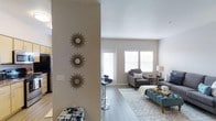 This single-floor, two-bedroom, two-bathroom residence offers a thoughtfully designed open-concept layout that seamlessly connects its living areas. The living room is a welcoming, light-filled space anchored by large windows dressed with white blinds that invite abundant natural light. Neutral-toned walls and light hardwood-style flooring run throughout, creating a clean, cohesive aesthetic that complements a variety of décor styles.

A partial wall with decorative accents elegantly separates the entryway from the main living area, adding architectural character while maintaining an open, airy feel. The transition between spaces is fluid, with a breakfast bar visible near the kitchen zone, providing a casual seating option ideal for everyday use.

The living area offers generous room for comfortable furnishings, as evidenced by the spacious seating arrangement visible in the image. Recessed and standard lighting fixtures provide warm illumination throughout. The overall ambiance is modern yet inviting, with clean lines, understated finishes, and a neutral palette that lends itself to effortless personalization. The two bedrooms and two full bathrooms provide practical, comfortable accommodations suited for a range of lifestyles.