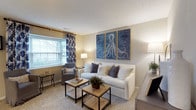 Located at 7999 Wildflower Ln in Westerville, Ohio, this two-story home offers a thoughtfully designed layout with comfortable and functional living spaces across both levels. The living room sets a welcoming tone with plush carpeting, neutral wall tones, and a textured ceiling that adds subtle character. A generously sized window dressed with decorative blue and white patterned curtains allows natural light to fill the room, creating a bright and airy atmosphere. The space is well-proportioned, comfortably accommodating a full seating arrangement centered around rustic wooden coffee tables. A striking tri-panel nautical wall art piece serves as a focal point, lending the room a coastal-inspired aesthetic that ties the décor together cohesively. The neutral color palette of beige, white, and gray throughout the living area creates a calm and inviting ambiance. The home's two-floor layout provides a natural separation between living and private spaces, offering both openness and functionality. Overall, the interior presents a clean, well-maintained environment with a balance of comfort and style, making it a welcoming residence in the Westerville community.