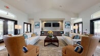 This well-appointed single-floor communal space is designed to impress, offering a seamless blend of sophisticated style and comfortable functionality across several distinct zones. The main lounge area commands attention with its soaring vaulted ceiling and a striking whitewashed brick accent wall, which serves as the backdrop for dual mounted flat-screen televisions flanked by dark wood cabinetry. Plush white sofas, warm cognac leather armchairs, and a substantial wood coffee table create an inviting and elegantly furnished gathering space.

Adjacent to the main lounge, a secondary seating area features wide-plank wood-look flooring, a bold navy velvet sofa set against a paneled accent wall with decorative sconce lighting, and a variety of eclectic seating arrangements that encourage both socializing and relaxed lounging.

A well-designed bar area offers a generous white countertop island with colorful upholstered barstools, illuminated by a row of handblown glass pendant lights. A whitewashed brick backsplash and open wood shelving complete the curated aesthetic behind the bar.

The entry corridor is bright and welcoming, featuring tall windows and sleek glass-paneled double doors that flood the space with natural light. Throughout, large-format tile and wood-look flooring, dark-framed windows, and carefully selected artwork contribute to a cohesive, modern design with a warm, inviting ambiance.
