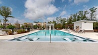 Located at 105 Ivy Green Way in Charleston, SC, this outdoor amenity space centers around a generously sized, sparkling swimming pool with crystal-clear turquoise water. The pool deck is thoughtfully designed to accommodate both relaxation and socializing, featuring built-in in-water loungers that allow residents to recline while partially submerged—ideal for warm South Carolina days. Surrounding the pool, a broad concrete deck provides ample space for rows of chaise lounges and seating arrangements, perfect for sunbathing or gathering with neighbors.

A modern, white pool house with large floor-to-ceiling windows serves as the architectural focal point of the space, offering interior access and a pass-through window along the exterior wall for added convenience. An additional pergola structure on the opposite end of the deck provides a shaded retreat for those seeking relief from direct sunlight. Brick accents and lush potted plantings add warmth and a touch of greenery to the hardscape surroundings.

The space is enclosed by a wrought iron fence, ensuring privacy and safety, while mature trees lining the perimeter frame open views of the surrounding Charleston sky. The overall ambiance is bright, clean, and resort-inspired, making this pool area a standout communal feature of the property.