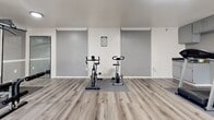 Located in Garden Grove, CA, this dedicated fitness room offers a well-organized workout space designed to support a variety of exercise routines. The room is finished with light-toned luxury vinyl plank flooring throughout, contributing to a clean, modern aesthetic that complements the neutral gray walls and white trim.

The gym is equipped with a full multi-station cable weight machine anchored in one section of the room, paired with a flat bench and additional free weights — ideal for strength training. A large mirror panel along one wall enhances the sense of space and supports form-checking during workouts. In the cardio zone, two treadmills and multiple stationary bikes — including a cycling-style spin bike — provide ample options for aerobic exercise.

Vertical blinds cover multiple large sliding glass door panels along the exterior wall, allowing natural light to filter in while maintaining privacy. A wall-mounted air conditioning unit ensures climate comfort year-round. Recessed and flush-mount ceiling lighting provides consistent illumination throughout the space.

A built-in kitchenette area along one wall features gray cabinetry with a dark countertop and a small sink, offering convenient access to water and storage. Crown molding accents the ceiling, adding a refined touch to this practical and fully functional fitness space.