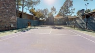 This single-story property offers a well-rounded combination of outdoor recreational amenities and functional interior living spaces. The centerpiece of the exterior is a full-sized tennis court with a light blue and green playing surface, complete with a regulation net and white court markings. The court is enclosed by chain-link fencing and is framed by mature pine and deciduous trees that provide natural shade and a scenic backdrop. Surrounding brick and stone-accented buildings contribute to the established, residential character of the setting.

Inside, the living area features dark hardwood-style flooring that extends throughout, creating a warm and cohesive aesthetic. A textured ceiling with a ceiling fan adds to the comfortable ambiance of the space. Neutral white walls provide a clean, versatile backdrop throughout the interior. A well-lit hallway lined with large windows dressed in white horizontal blinds allows natural light to fill the corridor, leading toward an exterior door. Recessed lighting and an emergency exit sign indicate thoughtfully integrated functional elements. Overall, the space balances practical amenities with a relaxed, inviting atmosphere suited for both activity and everyday living.