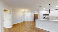 This thoughtfully designed single-floor residence showcases a clean, modern aesthetic with an open-concept floor plan that seamlessly connects the kitchen, dining, and living areas. Warm hardwood-style flooring flows consistently throughout, anchoring the bright, neutral color palette of soft grays and whites.

The kitchen is a standout feature, outfitted with crisp white shaker cabinetry, marble-look tile backsplash, and light quartz countertops that extend across a generous peninsula with an undermount sink and matte black faucet. Stainless steel appliances — including a gas range, microwave, and French door refrigerator — are complemented by striking black and gold cylindrical pendant lights that add a contemporary touch.

The open living and dining area benefits from abundant natural light streaming through sliding glass doors that lead to an outdoor balcony, providing pleasant exterior views. A bold sputnik-style chandelier with brass accents serves as a focal point in this shared space, while built-in open shelving adds both function and character near the entryway.

The bedroom is a serene retreat, featuring a large window with views, warm flooring, and a modern ring-style ceiling light fixture. The bathroom is finished with white cabinetry, quartz countertops, a large backlit vanity mirror, and crisp tile flooring, creating a clean and polished environment throughout.