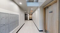The lobby at 4814 Interlake Ave N in Seattle, WA offers a streamlined, contemporary entry experience designed with both function and aesthetics in mind. The corridor-style layout is finished with light-toned large-format tile flooring and crisp white walls accented by dark baseboards, creating a clean, modern contrast throughout the space.

Natural light filters in through a skylight positioned near the entrance, complementing the recessed ceiling lighting that keeps the corridor well-illuminated. A glass entry door at one end provides a view to the exterior walkway and surrounding greenery, reinforcing a sense of openness upon arrival.

Resident mailboxes are mounted flush into the wall along one side of the corridor, keeping the design tidy and unobtrusive. Adjacent to these, a Luxer One automated package locker system provides a secure and convenient solution for parcel delivery. An elevator is accessible further along the corridor, flanked by dark-finished doors that contrast with the lighter surrounding finishes. A fire safety station is also conveniently located along the hallway.

The lobby transitions smoothly into the building's interior corridor, where wood-toned unit doors add warmth to the otherwise neutral palette. Overall, the space presents a well-organized, efficiently designed entry point that balances practicality with a polished, contemporary aesthetic.
