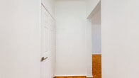 Located at 432 S New Hampshire Ave in the heart of Los Angeles, CA, this single-floor unit offers a well-organized layout that maximizes every square foot. The open-concept main area seamlessly combines the living room, dining space, and kitchen, creating a cohesive and functional environment. Warm parquet wood flooring extends throughout the living and dining areas, lending a classic character to the bright, white-walled interior. A ceiling fan anchors the living space, while a large picture window invites natural light and outdoor views. A built-in shelving unit with an integrated desk provides a practical workspace and display area, adding architectural interest to the main living zone. The kitchen is outfitted with white cabinetry, a gas range, full-size refrigerator, and a built-in range hood, with tile flooring that transitions cleanly from the parquet. A breakfast bar with seating connects the kitchen to the living area. The dining nook benefits from a window overlooking the street, keeping the space light-filled. The bathroom features a pedestal sink, toilet, a shower-tub combination, and tile flooring, all finished in a clean, minimalist white palette. Two hallways provide efficient circulation throughout the unit, contributing to a well-connected and easy-to-navigate floor plan.