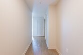 This contemporary single-floor residence showcases a thoughtfully designed layout with high-quality finishes and a clean, modern aesthetic. The kitchen is a standout feature, outfitted with high-gloss white upper cabinetry, warm wood-toned lower cabinets, and a teal subway tile backsplash. Stainless steel appliances — including a refrigerator, range, microwave, and dishwasher — complement the neutral stone countertops, creating a polished and functional cooking space. Light hardwood flooring flows throughout the main living areas, contributing to a bright and open atmosphere. The spacious living area benefits from track lighting and a seamless connection to the rest of the unit, with a sliding frosted door adding a modern architectural touch. A hallway with rich hardwood floors and louvered doors leads between spaces with clean sightlines. The bedroom features plush wall-to-wall carpeting and a floor-to-ceiling window that frames urban skyline views, flooding the room with natural light. The bathroom is generously proportioned, featuring a dual-sink vanity with stone countertops, warm wood cabinetry, a large wall mirror, and a combination tub and shower. Throughout the residence, neutral tones and contemporary detailing create a cohesive and refined living environment.