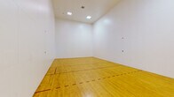 This dedicated indoor racquetball court is a purpose-built athletic facility designed to meet the demands of competitive and recreational play alike. The court features a classic hardwood floor finished in a warm golden tone, with clearly marked red boundary and service lines that define the regulation playing area. The expansive white walls on all sides provide a clean, unobstructed playing surface, essential for the fast-paced nature of racquetball. The ceiling rises to a generous height, allowing full freedom of play and accommodating the dynamic movement of the game. Overhead, recessed flush-mounted light panels provide even, consistent illumination throughout the court without creating glare or shadows. Entry to the court is provided through a glass door fitted with sturdy hardware, offering a view to an outdoor walkway and landscaped surroundings with tropical palms visible through the frame. A built-in utility access panel is visible along the back wall near the entrance. The overall environment is clean, minimal, and purpose-driven, with every element designed to support an optimal playing experience. This single-floor facility delivers a professional-grade recreational space ideal for fitness and sport enthusiasts.