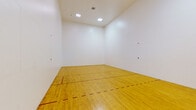 This dedicated indoor racquetball court is a purpose-built athletic facility designed to meet the demands of competitive and recreational play alike. The court features a classic hardwood floor finished in a warm golden tone, with clearly marked red boundary and service lines that define the regulation playing area. The expansive white walls on all sides provide a clean, unobstructed playing surface, essential for the fast-paced nature of racquetball. The ceiling rises to a generous height, allowing full freedom of play and accommodating the dynamic movement of the game. Overhead, recessed flush-mounted light panels provide even, consistent illumination throughout the court without creating glare or shadows. Entry to the court is provided through a glass door fitted with sturdy hardware, offering a view to an outdoor walkway and landscaped surroundings with tropical palms visible through the frame. A built-in utility access panel is visible along the back wall near the entrance. The overall environment is clean, minimal, and purpose-driven, with every element designed to support an optimal playing experience. This single-floor facility delivers a professional-grade recreational space ideal for fitness and sport enthusiasts.